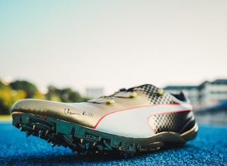 A track and field sports shoe is positioned on a blue running track, with soft sunlight illuminating its design. The shoe features sharp cleats underneath, a sleek metallic finish, and a signature inscribed on the side. Blurred buildings and greenery are visible in the background.