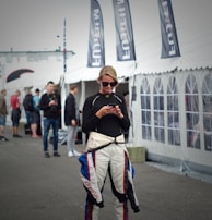 A person wearing a racing suit is standing on a hoverboard while using a smartphone. Several black flags with white lettering are visible behind, as well as a large white tent. Other people are in the background, suggesting a busy outdoor setting.