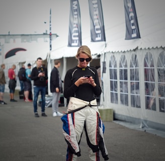 A person wearing a racing suit is standing on a hoverboard while using a smartphone. Several black flags with white lettering are visible behind, as well as a large white tent. Other people are in the background, suggesting a busy outdoor setting.