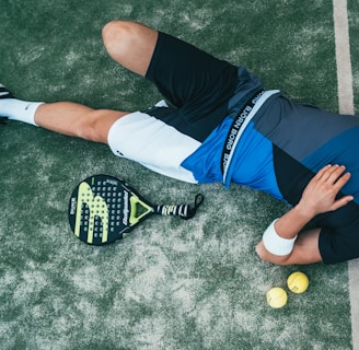 A person is lying on the ground, wearing athletic clothing that includes a blue and black top and white shorts. Nearby, there is a paddle racket and two yellow balls on a textured surface, which looks like a sports court. The person is wearing black sneakers and has a wristband on their arm.