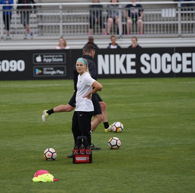 A person in sports attire stands on a soccer field, surrounded by several soccer balls and a carrier with bottles. The field is bordered by advertisements for Nike Soccer and app stores. Several people sit in bleachers in the background.