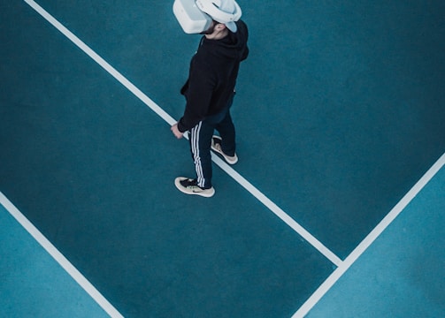 A person wearing a virtual reality headset stands on a large, empty sport court with distinct white lines on the floor. The dark clothing contrasts with the bright headset.