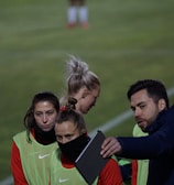 A group of individuals on a sports field are wearing green vests over red attire. One person is holding a tablet and seems to be discussing or showing something to the others. The atmosphere suggests a sports event, likely soccer, indicated by their clothing and the setting. The background is a grassy field.