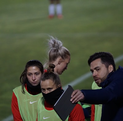 A group of individuals on a sports field are wearing green vests over red attire. One person is holding a tablet and seems to be discussing or showing something to the others. The atmosphere suggests a sports event, likely soccer, indicated by their clothing and the setting. The background is a grassy field.