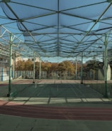 A spacious indoor sports facility with a tennis court and running tracks. The court is surrounded by high nets and tall, green metal structures supporting a roof. The area is well-lit with natural light filtering in through a transparent ceiling. In the background, there are trees with autumn foliage and several residential buildings.