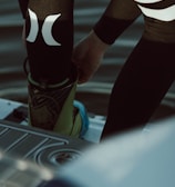 A person is putting on or adjusting water sports gear, standing on a platform above a body of water. The lower part of their body is visible, dressed in a black wetsuit with prominent white logos. Their footwear appears specialized, suggesting activities like wakeboarding or water skiing.