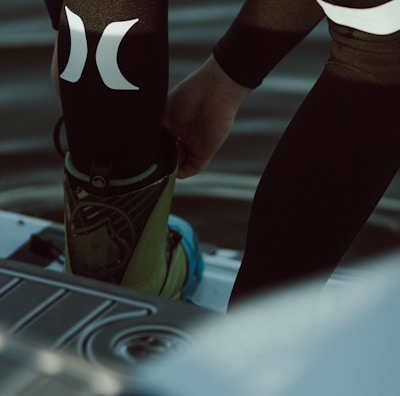 A person is putting on or adjusting water sports gear, standing on a platform above a body of water. The lower part of their body is visible, dressed in a black wetsuit with prominent white logos. Their footwear appears specialized, suggesting activities like wakeboarding or water skiing.
