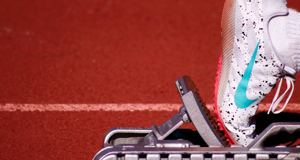 A close-up of a white athletic shoe with colorful speckles and a blue swoosh logo, resting on a starting block on a red track field. The shoe is positioned as if ready for a race, with a visible foot wearing a white sock inside the shoe.