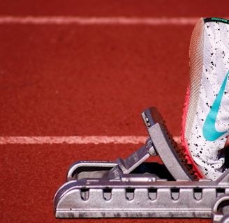 A close-up of a white athletic shoe with colorful speckles and a blue swoosh logo, resting on a starting block on a red track field. The shoe is positioned as if ready for a race, with a visible foot wearing a white sock inside the shoe.