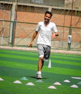 A person in sports clothing is running on a green artificial turf with white and red cones arranged in a pattern. The individual appears to be participating in a sports drill or exercise. The background includes a brick wall and netting consistent with an outdoor sports facility.