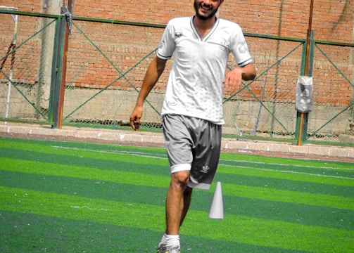 A person in sports clothing is running on a green artificial turf with white and red cones arranged in a pattern. The individual appears to be participating in a sports drill or exercise. The background includes a brick wall and netting consistent with an outdoor sports facility.