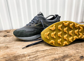 A pair of dark-colored athletic shoes placed on a wooden surface, with one shoe laying on its side to display the sole. The shoes have a sleek design with a mix of suede and mesh materials, featuring a rubber sole with unique tread patterns for grip.