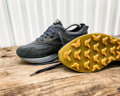 A pair of dark-colored athletic shoes placed on a wooden surface, with one shoe laying on its side to display the sole. The shoes have a sleek design with a mix of suede and mesh materials, featuring a rubber sole with unique tread patterns for grip.