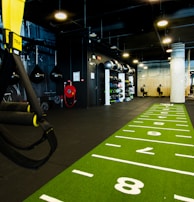 A modern gym space featuring a green track laid out on the floor with numbered lanes. TRX suspension trainers hang prominently in the foreground. The gym is equipped with various exercise equipment, including kettlebells and punching bags. In the background, there is a FIT sign near a reception desk under a low-ceilinged industrial design.