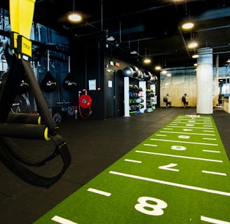 A modern gym space featuring a green track laid out on the floor with numbered lanes. TRX suspension trainers hang prominently in the foreground. The gym is equipped with various exercise equipment, including kettlebells and punching bags. In the background, there is a FIT sign near a reception desk under a low-ceilinged industrial design.