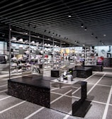 A modern sneaker store with sleek black shelving displaying various athletic shoes and sports apparel. Central seating areas and spotlights provide a contemporary retail ambiance. The signage prominently features a famous sportswear slogan in the background.