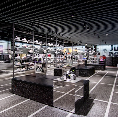 A modern sneaker store with sleek black shelving displaying various athletic shoes and sports apparel. Central seating areas and spotlights provide a contemporary retail ambiance. The signage prominently features a famous sportswear slogan in the background.