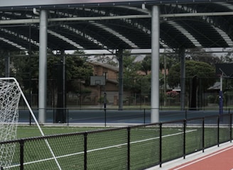 An outdoor sports facility featuring a running track and a soccer goal. The track is red, surrounded by a black metal fence, and the field is covered with artificial green turf. In the background, a basketball court is visible under a covered roof with metal supports. The sky is overcast, adding a moody atmosphere.
