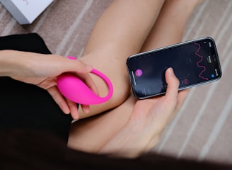 A person holds a bright pink wearable device in one hand while interacting with a smartphone in the other hand. The smartphone screen displays a graph with lines and numerical data, suggesting a tracking or monitoring application. The background consists of a soft, neutral-toned surface with a partial view of a box.