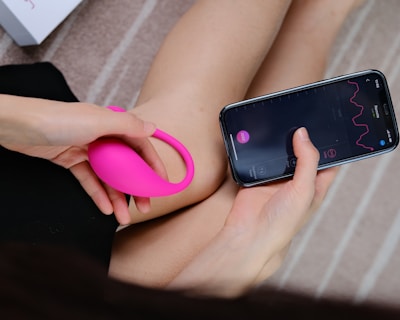 A person holds a bright pink wearable device in one hand while interacting with a smartphone in the other hand. The smartphone screen displays a graph with lines and numerical data, suggesting a tracking or monitoring application. The background consists of a soft, neutral-toned surface with a partial view of a box.