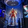 A high-tech suit displayed in a futuristic setting, illuminated by focused lighting. The surroundings include digital screens showing technical data and designs related to the suit. The environment appears to be a lab or exhibit with metallic and technological elements.