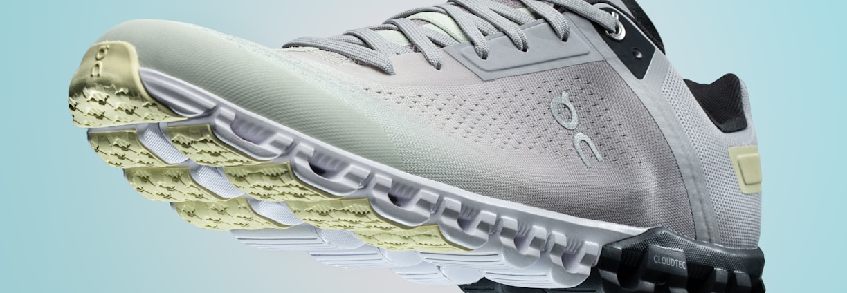 A single sports shoe is prominently displayed against a seamless, light turquoise background. The shoe features a sleek design with a gradient of light gray to dark gray tones, and is equipped with a distinctive CloudTec sole for cushioning. The branding logo is subtly visible on the side.