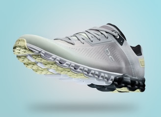 A single sports shoe is prominently displayed against a seamless, light turquoise background. The shoe features a sleek design with a gradient of light gray to dark gray tones, and is equipped with a distinctive CloudTec sole for cushioning. The branding logo is subtly visible on the side.