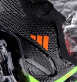 A close-up view of a sleek black sports shoe with a textured surface and an orange logo. The sole has a shiny finish with molded studs, and there is a neon green accent at the bottom.