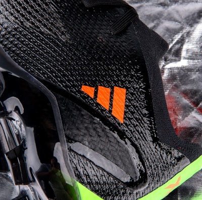 A close-up view of a sleek black sports shoe with a textured surface and an orange logo. The sole has a shiny finish with molded studs, and there is a neon green accent at the bottom.