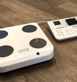 A digital body composition analyzer featuring a square platform with four circular metal sensors and an attached console with a display and various buttons. Both components are white with a sleek, modern design, placed on a wooden floor.