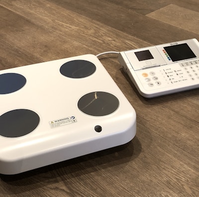 A digital body composition analyzer featuring a square platform with four circular metal sensors and an attached console with a display and various buttons. Both components are white with a sleek, modern design, placed on a wooden floor.
